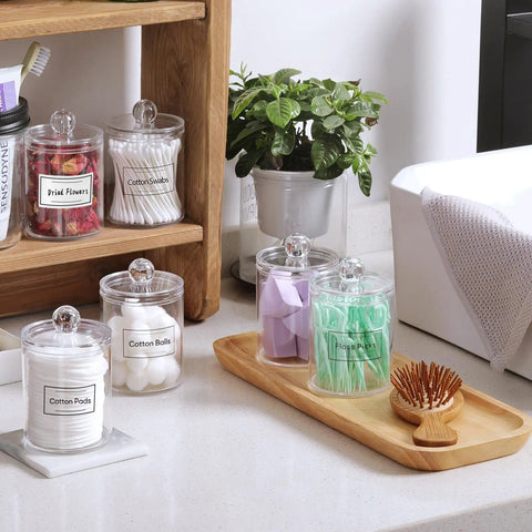 Super Practical Four Pack Clear Acrylic Qtip Holder Dispensers With Lids For Organizing Cotton Swabs Balls Pads And Floss Picks Comes With Labels In Two Sizes Twelve Ounce And Ten Ounce on bathroom counter.