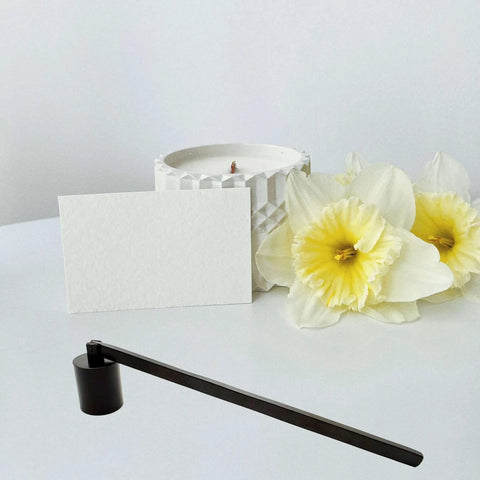 Sleek Black Stainless Steel Candle Snuffer With Rotating Swivel Bell And Ergonomic Grip Handle For Smooth Flame Control and Deep Jar Reach next to a white candle and yellow flowers.