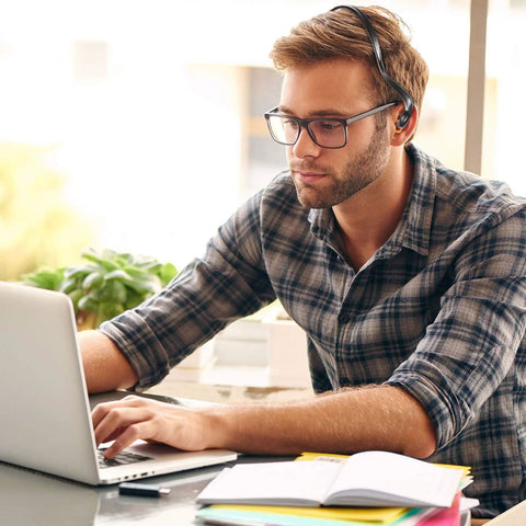 Man wearing flannel shirt and glasses using laptop at desk with headset on, focused on work