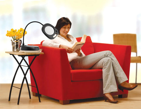 Woman reading next to Go-To LED Floor Lamp And Table Lamp With 5-Inch Magnifier on side table in living room setting.