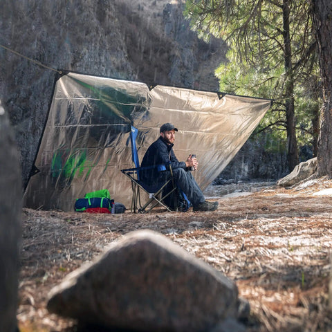 Person sitting under Rugged Survival Tarp With Insulated Reflective Mylar Layer For All Weather Camping, woodland camo setting.