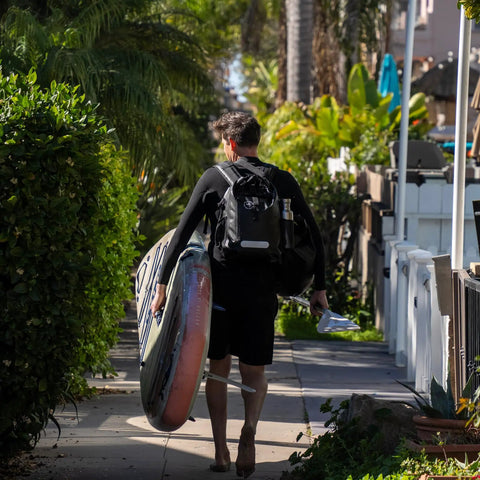 Man walking to beach carrying surfboard and wearing 25L Waterproof Surf Backpack For Adventure With Roll Top Closure Keeps Gear Dry