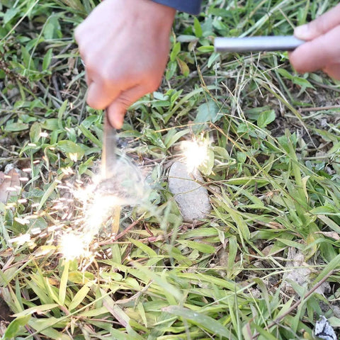Person using Survival Fire Starter Kit With Drilled Ferrocerium Rod Half Inch By Six Inch Striker to create sparks on grass.
