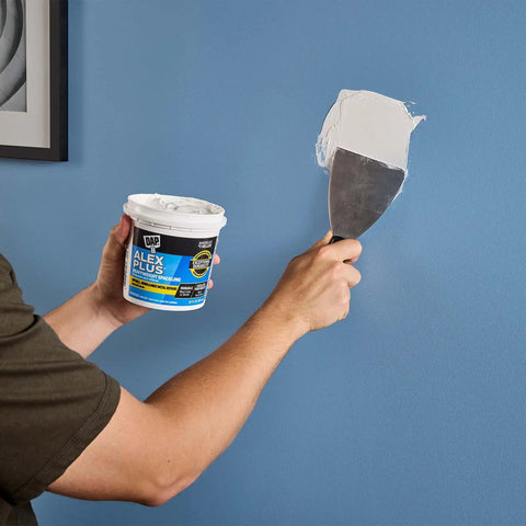 Person applying Alex Plus Spackling White 32 Oz Quick Dry Patch For Drywall Wood And Metal No Sag No Shrink on a blue wall.