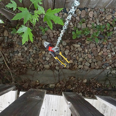 Top view of pliers with yellow handles hanging from a chain, outdoor setting with rocks and leaves visible.