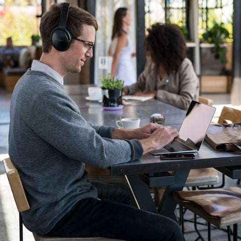 Man using Premium Wireless Bluetooth Headphones Delivering Game Changing Noise Cancellation And Seamless Alexa Access For Lock In Music Vibes And Zero Stress Comfort All Day Long In Black while working on a laptop at a cafe.