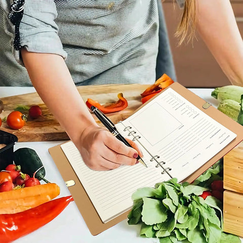 A person using the Leather Recipe Book With Index Pages And Templates, The Ultimate Recipe Notebook For Recording Your Own Dishes, Blank Cookbook To Organize Flavorful Creations, Waterproof Cover For Everyday Kitchen Use, alongside fresh vegetables in a kitchen.