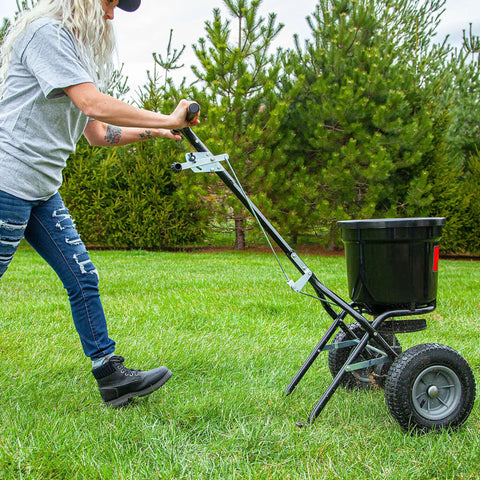 Person using 50 Lb Capacity Push Broadcast Spreader For Lawns And Gardens, Easy Push Handle, Durable Heavy Duty Frame, Adjustable Spreading Rate