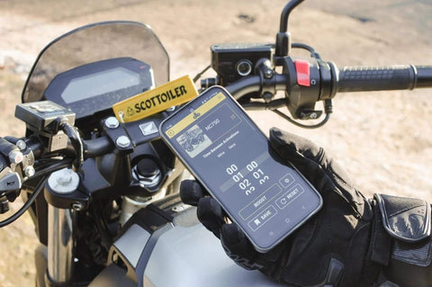 Gloved hand holding smartphone with Essentials Micro XSystem 3.0 Plus Electronic Automatic Motorcycle Chain Lubrication System app on bike